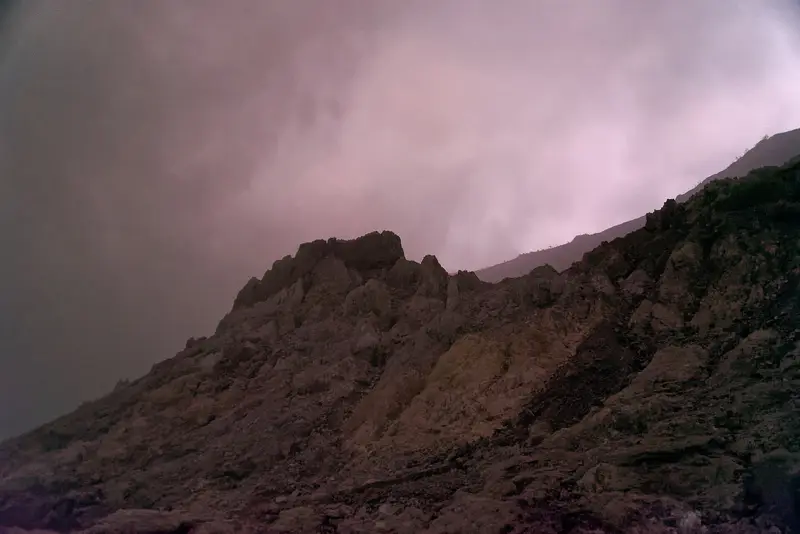 Rocky inner wall of Kawah Ijen crater under pink-lit gas clouds before sunrise